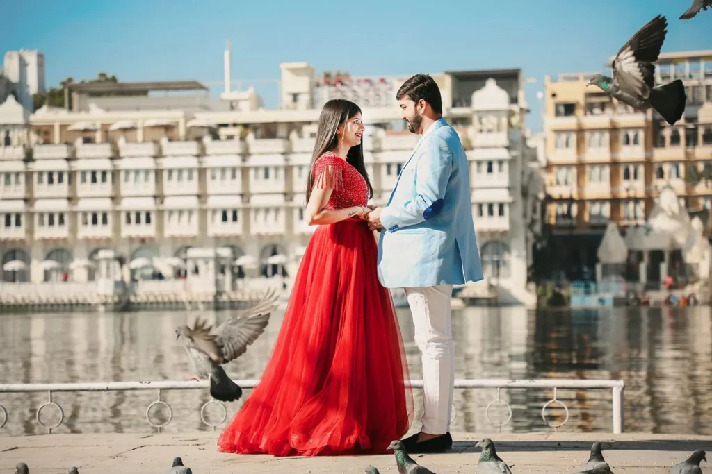 Couple in a red gown and a light blue blazer facing each other by a lake with pigeons flying around