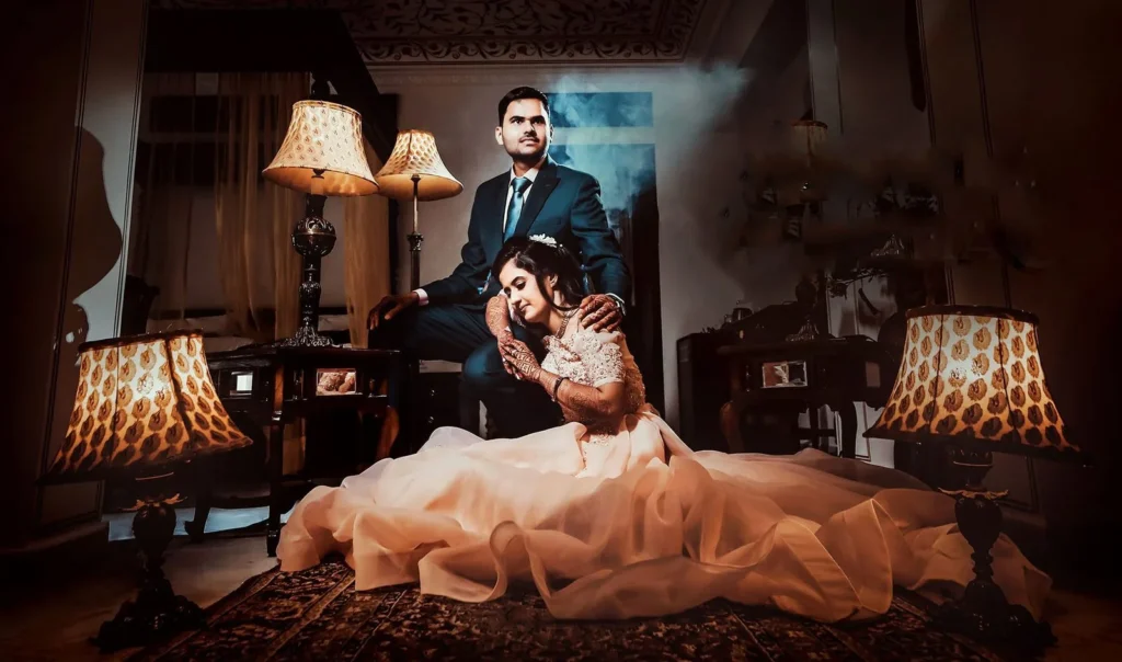 Groom standing and bride seated in a flowing gown in a lamp-lit vintage room, a wedding couple portrait