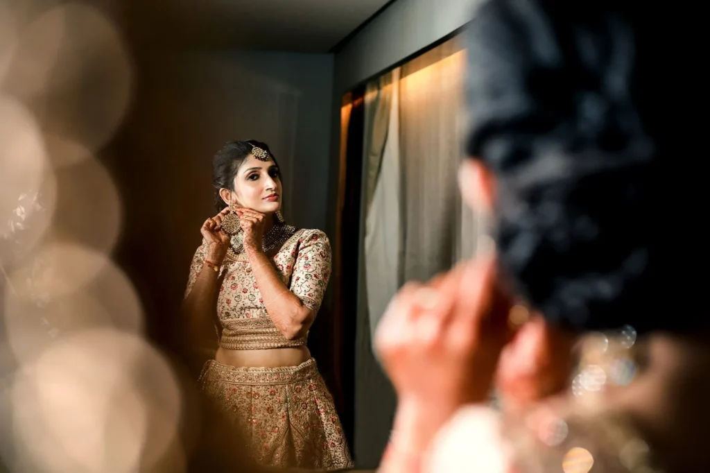 Bride in a beige embroidered lehenga adjusting her earring in front of a mirror before the wedding