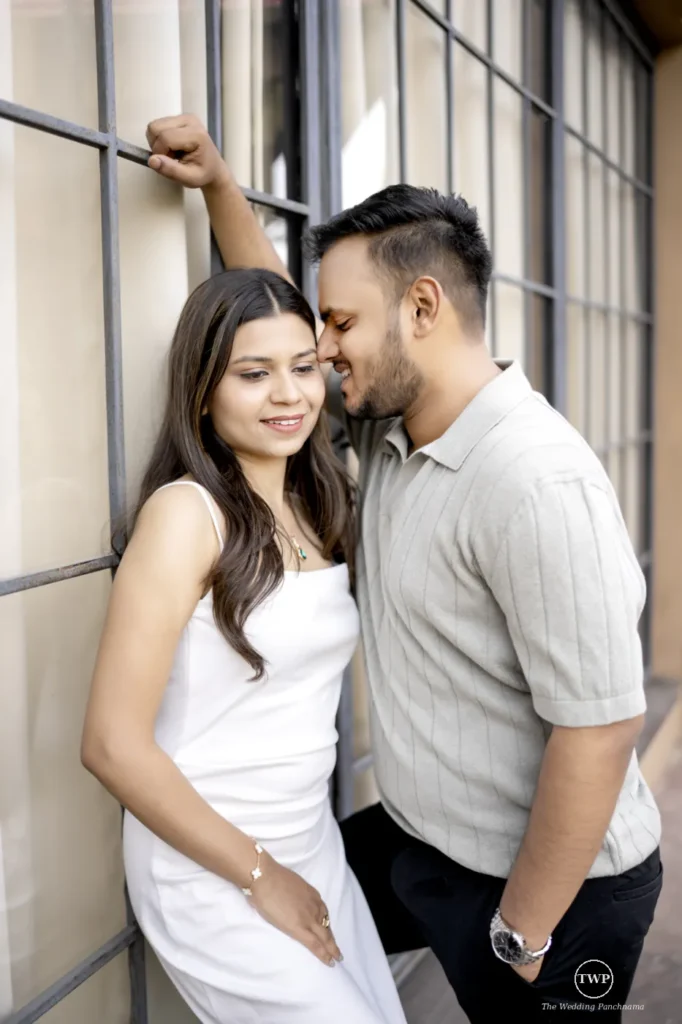A couple standing close by a large gridded window at home, the woman in a white dress smiling as the man leans in toward her cheek
