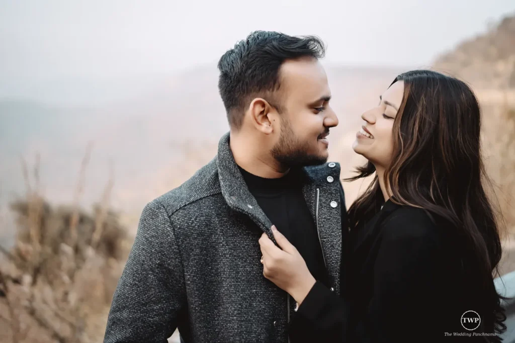 A couple smiling at each other on a hillside at golden hour, the woman in black holding the man's jacket collar