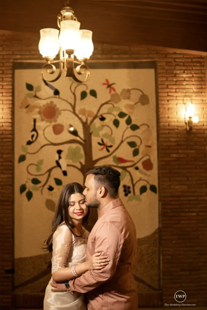 A couple sharing a quiet embrace under a chandelier, the man in a peach shirt kissing the smiling woman on the temple against a painted tree wall