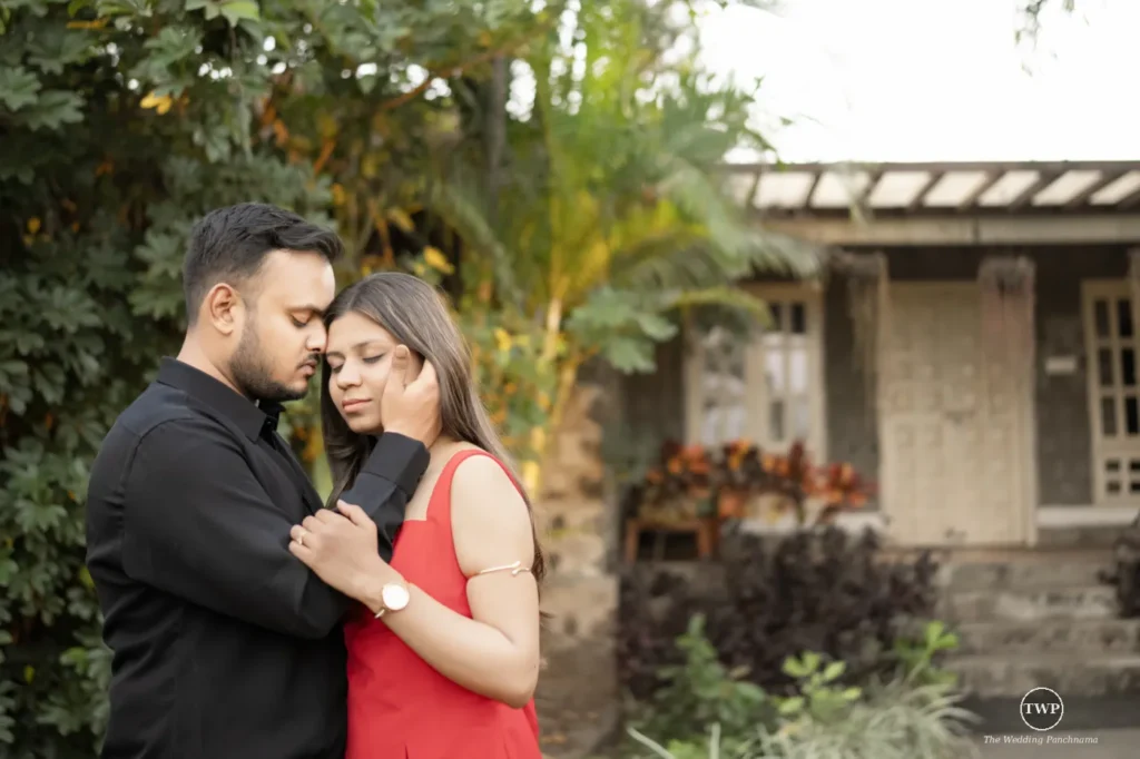 A couple standing forehead to forehead in a garden beside a stone cottage, the woman in a red dress and the man in black