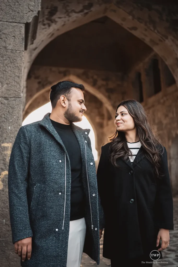 A couple in dark winter coats smiling at each other under a weathered stone archway