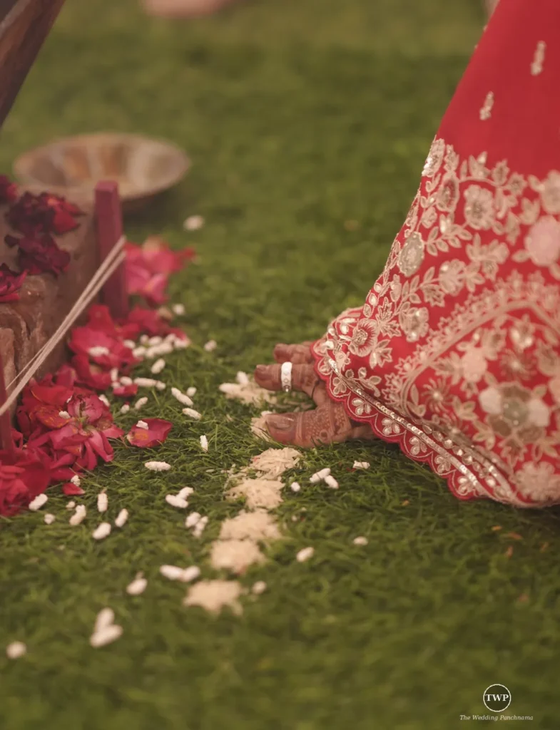 An embroidered red bridal lehenga hem beside a mehendi-decorated foot, with rose petals and rice scattered on green grass