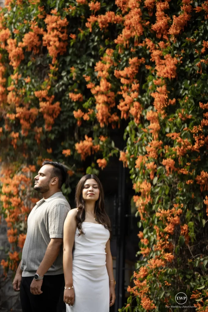 Couple standing back to back before a wall of orange flowers on their floral pre-wedding shoot near Indore
