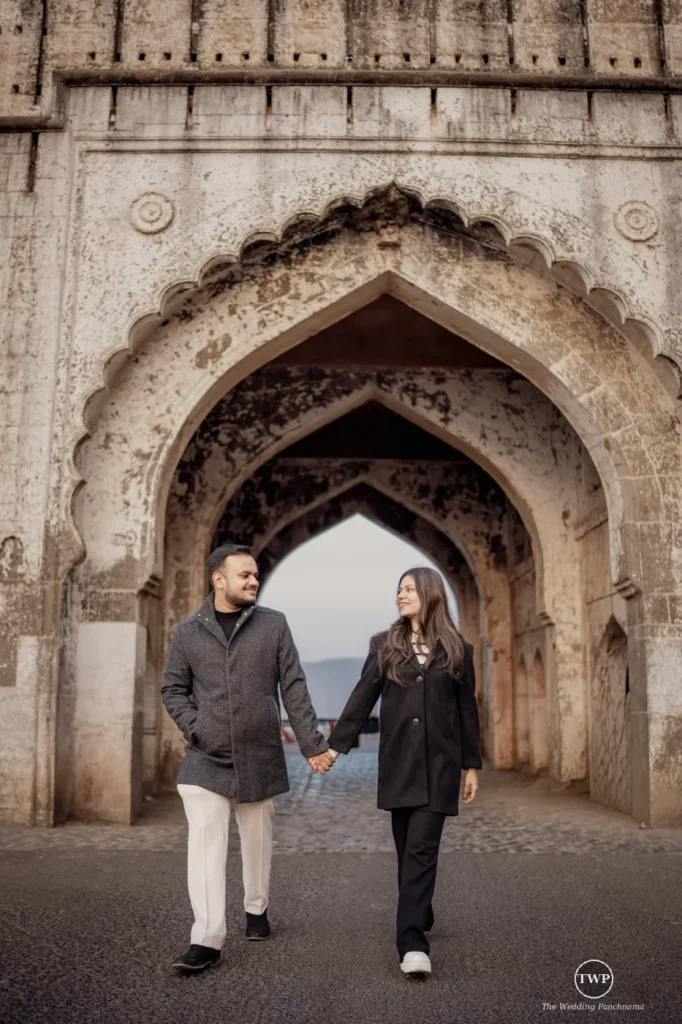 A couple holding hands and walking under a carved stone archway with a lake and hills behind them