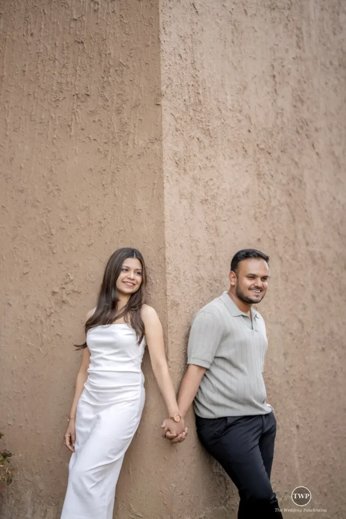 A couple holding hands and leaning against a textured mud-plaster wall, the woman in a white dress and the man in a grey polo shirt