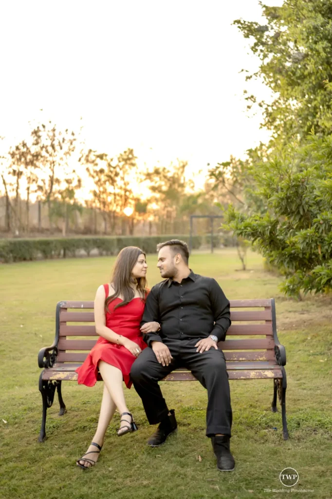A couple sitting close together on a wooden park bench at sunset, the woman in a red dress and the man in black